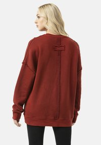 Oversized rode sweatshirt met versleten randen, gestructureerde stof en een centrale naad. Heeft afgezakte schouders en lange mouwen.
