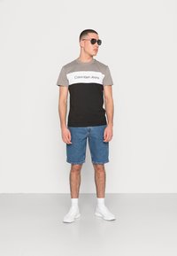 Camisa de color bloqueada en negro, gris y blanco con el logo "Calvin Klein Jeans", combinada con un short de denim y zapatillas blancas. El modelo lleva gafas de sol.
