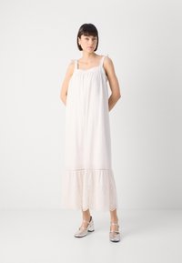 Vestido sin mangas de color rosa claro, con tirantes con volantes y dobladillo bordado. La tela suave cae hasta la mitad de la pantorrilla. Zapatos de tacón bloque plateados.