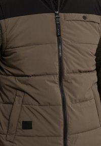Olivgrüne Steppjacke mit schwarzem Oberteil, Reißverschluss und Seitentaschen. Verfügt über eine weiche Textur und ein rechteckiges Stoffetikett.