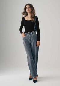 Sort langærmet top, høj talje grå denim jeans og sorte spidse sko. Model står foran en neutral baggrund med hænderne på hofterne.