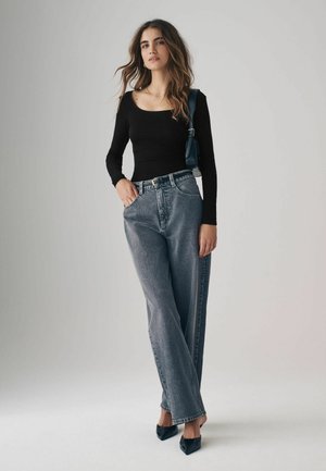 Maglia nera a maniche lunghe, jeans grigi a vita alta, e scarpe nere con punta. Il modello è in piedi su uno sfondo neutro, con le mani sui fianchi.