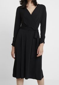 Svart omlottklänning med långa ärmar, djup V-ringning, självknut i midjan och svängande kjol. Tillverkad av mjuk, slät tyg med en bekväm passform.