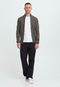 Gestrickte Zip-up-Cardigan in Dunkelgrau, mit hohem Kragen und gerippten Bündchen, getragen über einem weißen T-Shirt und kombiniert mit schwarzen Hosen und weißen Turnschuhen.