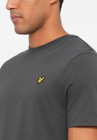 Close-up de um homem a usar uma t-shirt de manga curta cinza escuro com um pequeno logótipo de um pássaro amarelo no lado esquerdo do peito.
