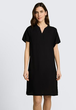 Kobieta z brązowymi włosami do ramion, ubrana w prostą czarną sukienkę do kolan z krótkimi rękawami i dekoltem w serek, stojąca na jednolitym tle.