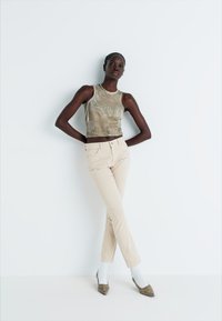 Beige slim-fit bikses apvienotas ar gaiši pelēku īsu topiņu. Modelis stāv ar vienu roku uz gurniem, valkājot baltas zeķes un pelēkus papēžus.