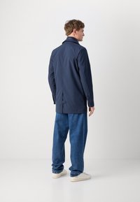 Giacca impermeabile blu marina con colletto alto, ventile posteriore e dettagli con bottoni, abbinata a pantaloni di jeans blu larghi e sneakers bianche.