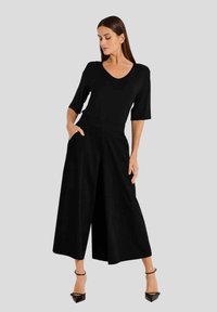 Zwarte V-hals top met korte mouwen, gecombineerd met wijdvallende zwarte broek. De broek heeft zijzakken. Model draagt zwarte hakken.