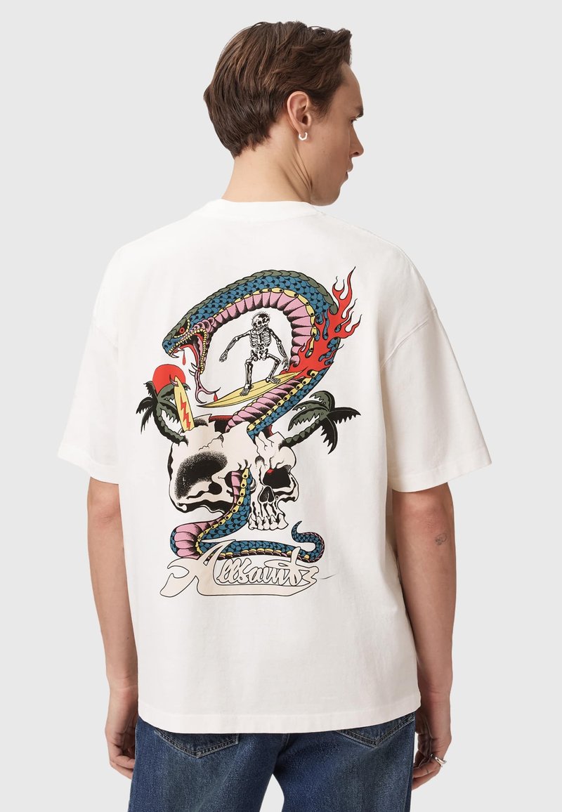 Mann trägt weißes T-Shirt mit farbenfrohem Motiv eines Skeletts, das auf einer Schlange surft, die um Totenköpfe und Palmen auf dem Rücken gewickelt ist.