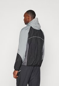 Veste coupe-vent colorée en blocs avec un design gris et noir, capuche réglable, poignets élastiques et accents réfléchissants dans le dos.