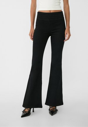 Femme portant un pantalon noir évasé taille haute et des talons noirs pointus avec des clous, associé à un crop top blanc.