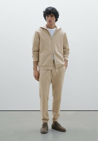 Beige hoodie met een ritssluiting aan de voorkant, zijzakken en geribde manchetten, gecombineerd met bijpassende taperende broek en grijze veterschoenen.