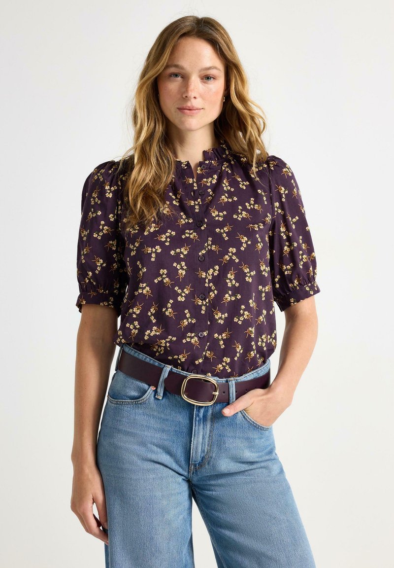 Lilla blomsterbluse med puffærmer, knaplukning foran og flæsekrave. Kombination med højtaljede lyseblå jeans og et mørkt bælte.