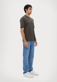 Camiseta de color verde oliva oscuro con mangas cortas, que presenta una impresión de logo en la parte delantera. Llevada con unos jeans azul claro y zapatillas blancas.