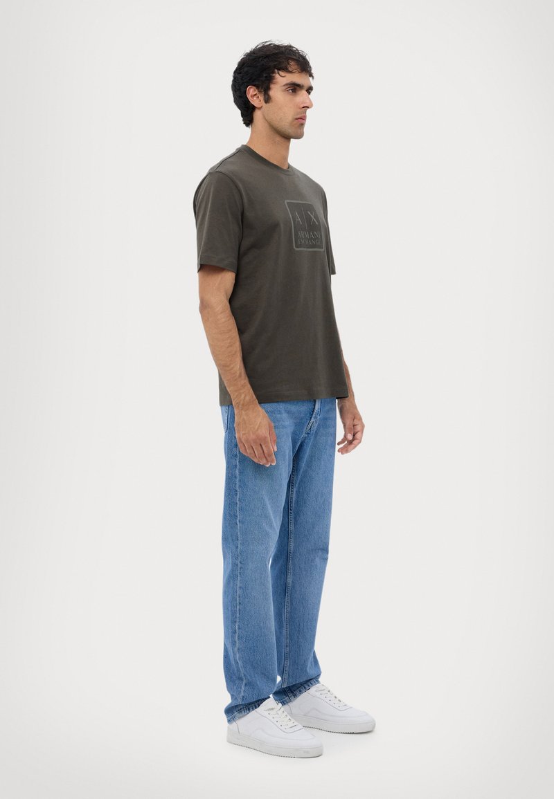 Camiseta de color verde oliva oscuro con mangas cortas, que presenta una impresión de logo en la parte delantera. Llevada con unos jeans azul claro y zapatillas blancas.