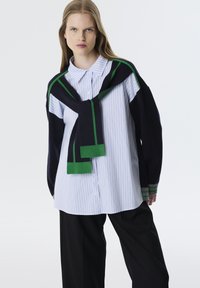 Gestreepte blauw-witte button-up shirt, gelaagd met een marineblauwe trui met groene accenten, gecombineerd met zwarte wijde broek.