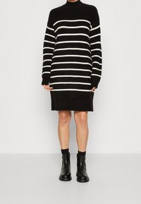 Robe tricotée à rayures noires et blanches avec un col montant et des manches longues, présentant une coupe droite et une texture douce. Portée avec des bottines noires.