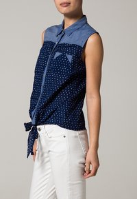 Top senza maniche di colore blu navy con colletto in denim, con pois bianchi sul corpo e un nodo legato in vita. Abbinato a jeans bianchi.