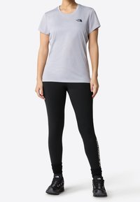 Jasnoszara sportowa koszulka z krótkim rękawem z czarnym logo, zestawiona z czarnymi legginsami z białym logo po boku, noszona z czarnymi butami.