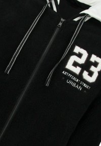 Zwarte fleece hoodie met ritssluiting, voorzien van witte geborduurde cijfers "23" en tekst "ADTTFERENT STREET URBAN", met gestreepte trekkoorden.