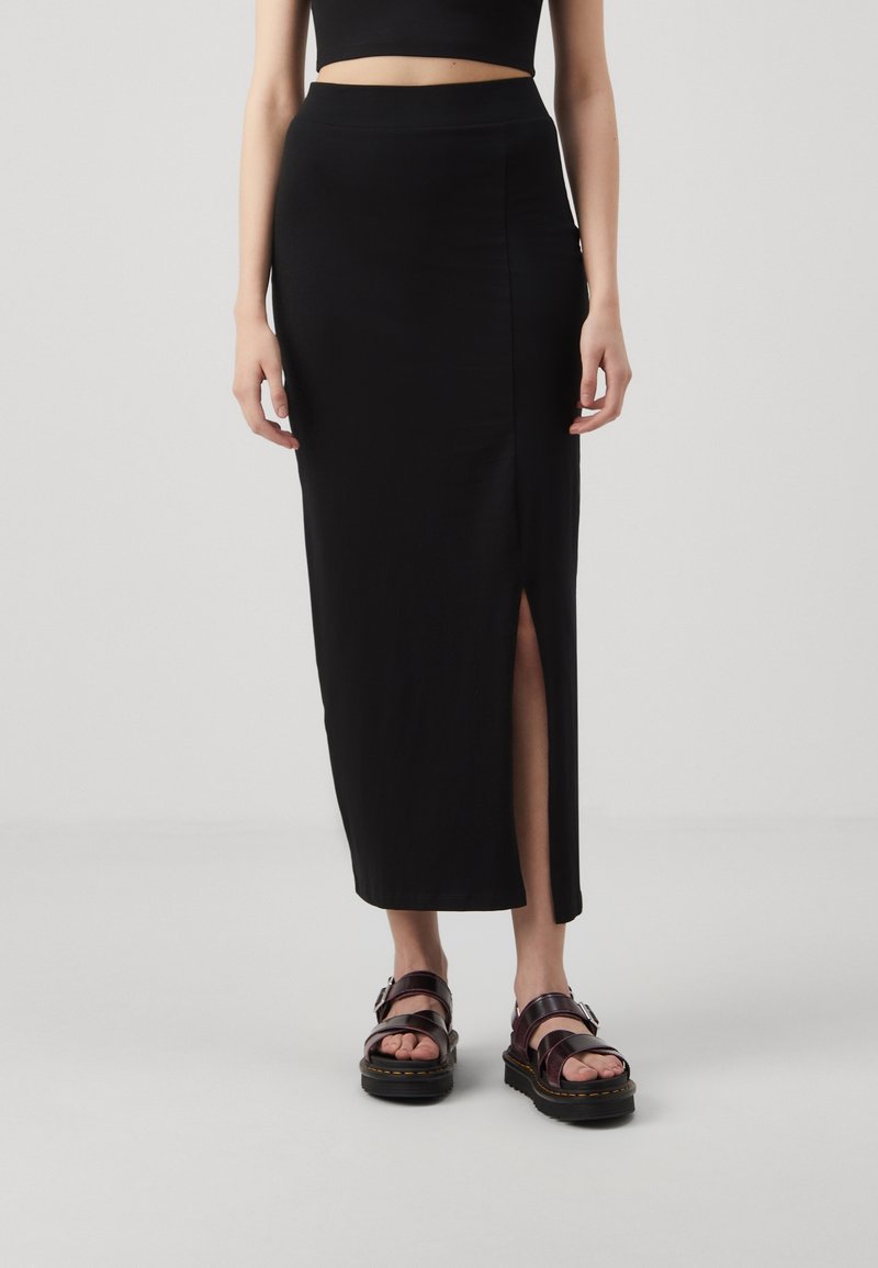 Femme portant une jupe midi noire avec une fente latérale, un crop top noir et des sandales épaisses marron foncé, debout sur un fond clair uni.