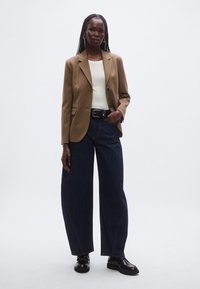 Pruun bleiser valge topi peal, sobitatud laia lõikega tumedate teksapükstega. Must rihm ja kingad täiendavad välimust, millel on sile tekstuur.