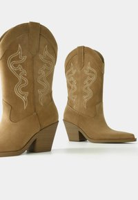 Beżowe kowbojki zamszowe z białymi przeszyciami na cholewce, spiczastym noskiem i słupkowym obcasem. Tekstura jest miękka i matowa.