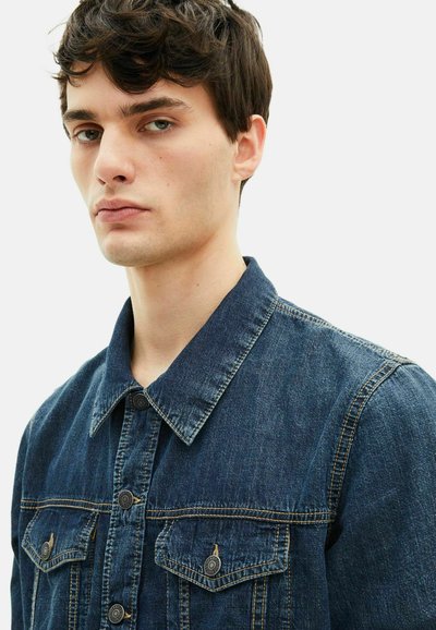 Jeansowa kurtka w ciemnym niebieskim kolorze z klasycznym kołnierzem, dwoma przednimi kieszeniami i metalowymi guzikami. Materiał jest lekko teksturowany i trwały.