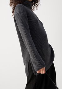 Šedý pletený svetr s rolákem a texturovaným vzorem, bočními rozparky a volným střihem, doplněný černými kalhotami, držený tenkým popruhem.