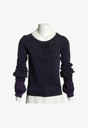 Pull bleu marine à manches longues avec des détails froncés au niveau de la poitrine et des poignets, superposé sur des manches bouffantes violet de chemise sur un mannequin blanc.