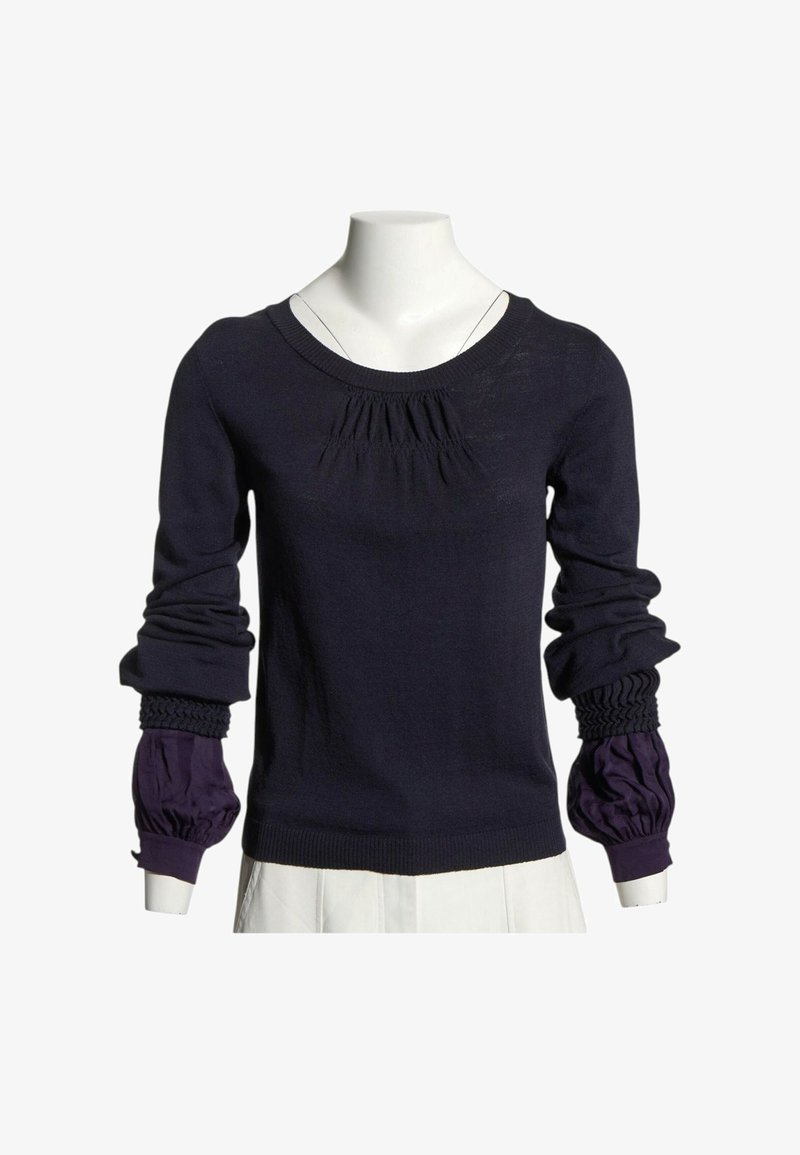Pull bleu marine à manches longues avec des détails froncés au niveau de la poitrine et des poignets, superposé sur des manches bouffantes violet de chemise sur un mannequin blanc.
