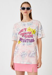 T-shirt med tie-dye i pastelfarver med korte ærmer, der har fed tekst "IGNORER DIN POTENTIALE IKKE" og gule blomsterdetaljer.