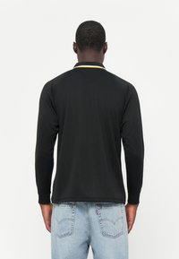 Polo shirt nera a maniche lunghe con una striscia del colletto gialla e bianca, caratterizzata da una texture liscia e una vestibilità comoda, indossata con jeans azzurri chiari.