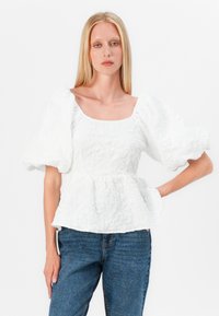 Blusa peplum bianca con maniche corte e voluminosi, tessuto texture, scollo quadrato e vita aderente, abbinata a jeans blu.
