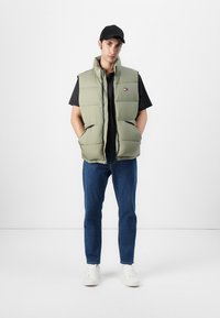 Olive green puffer vest with a zip front and two pockets, worn over a black t-shirt. Paired with blue jeans and white sneakers. Black cap.
