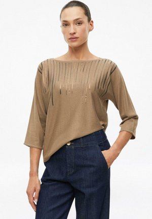 Frau trägt ein taupefarbenes Stricktop mit vertikalen Pailletten-Details und dunkelblaue High-Waist-Jeans, eine Hand in der Tasche, vor weißem Hintergrund.