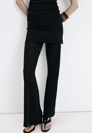 Kobieta w czarnych rozkloszowanych spodniach, czarnym tunikowym topie, czarnych sandałach na rzemykach i bransoletkach, stojąca przy białej ścianie.