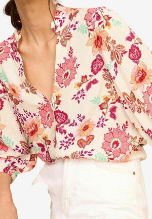 Blouse florale en tissu crème ornée de fleurs roses, rouges et jaunes vives, avec un col en V et des manches bouffantes. Associée à un pantalon blanc.