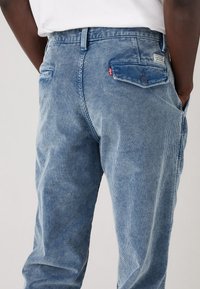 Blauwe corduroy broek met verticale strepen, voorzien van een achterzak met knoopsluiting en een klein rood logotag.