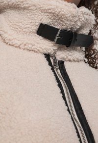 Chaqueta de forro polar con un tejido crema texturizado, cuello con correa de cuero negro y cierre de cremallera plateado a lo largo de una costura negra en contraste.
