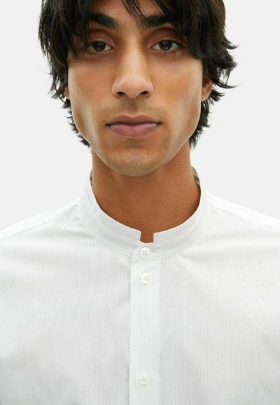 Jongere met een gemiddelde huidtint en donker haar draagt een witte, geknoopte blouse met een mandarinenkraag tegen een effen achtergrond.