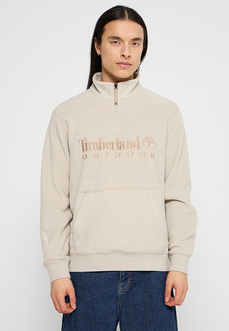 Pullover in pile beige con colletto alto, dotato di zip frontale e grande logo ricamato "Timberland OUTDOOR". Include una tasca frontale.