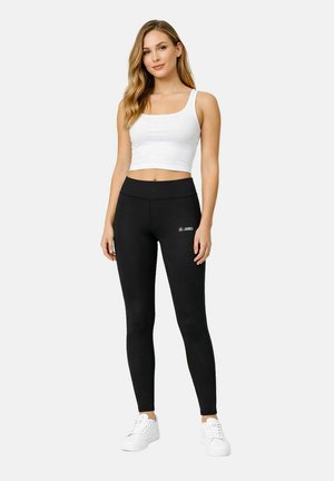 Donna in piedi che indossa un top bianco corto, leggings neri con logo Jako e scarpe da ginnastica bianche su uno sfondo chiaro uniforme.