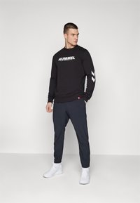 Svart langermet sweatshirt med "HUMMEL"-logo på fronten og hvit chevron-design på ermene, kombinert med marineblå smale bukser og hvite sneakers.