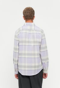 Hombre de espaldas vestido con una camisa de cuadros lila y gris de manga larga abotonada y pantalones gris oscuro, contra un fondo liso.