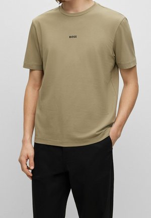 Personne portant un t-shirt marron clair avec le texte "BOSS" sur la poitrine et un pantalon noir, debout avec une main dans la poche.