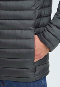 Puffjakke i mørkegrå med quiltet design. Har lynlåslommer på siden og struktureret stof. Let skinnende med en tætsiddende stil.