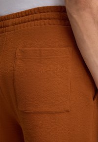 Roestkleurige joggingbroek van een textuurstof, met een open zak op de achterkant en een elastische tailleband.