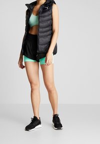 Svart pufferväst med huva, matchad med en ljusgrön sport-bh och svarta atletiska shorts. Svarta sneakers fulländar outfiten.